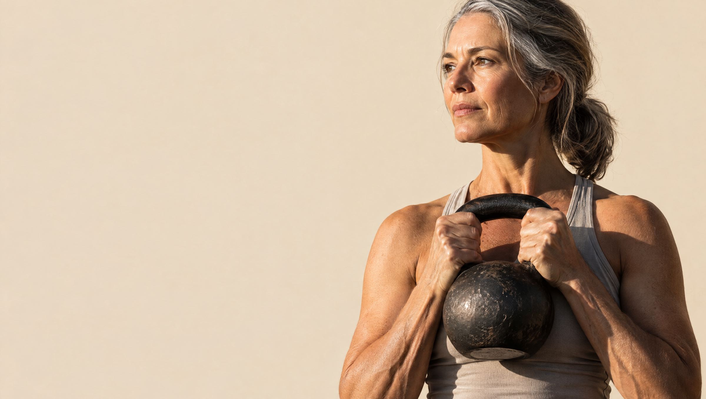 Mulher de cabelo grisalho segura um kettlebell na altura do peito, olhando para o lado, sob luz quente.